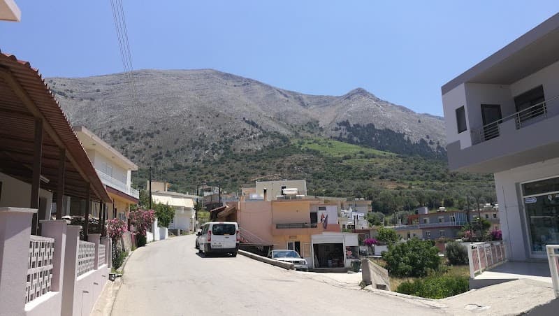 A serene village street in Greece features charming houses with balconies, blooming flowers, and a backdrop of lush mountains under a clear blue sky, ideal for travelers seeking a peaceful retreat.