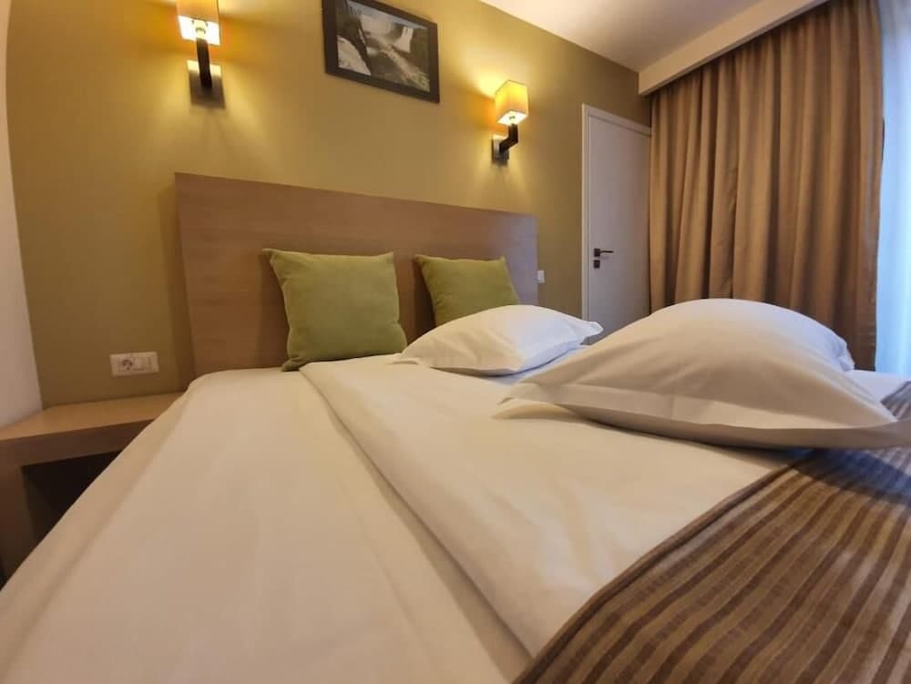 A cozy hotel room features a neatly made bed with white linens and green pillows, warm yellow walls, wall-mounted lamps, and a framed picture, ideal for travelers seeking comfort and relaxation.