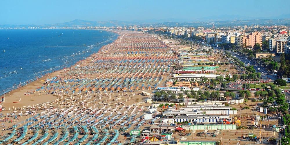 A vibrant coastal cityscape unfolds with a bustling beach lined with colorful umbrellas and sunbeds, adjacent to urban buildings under a clear blue sky, ideal for summer vacation planning.