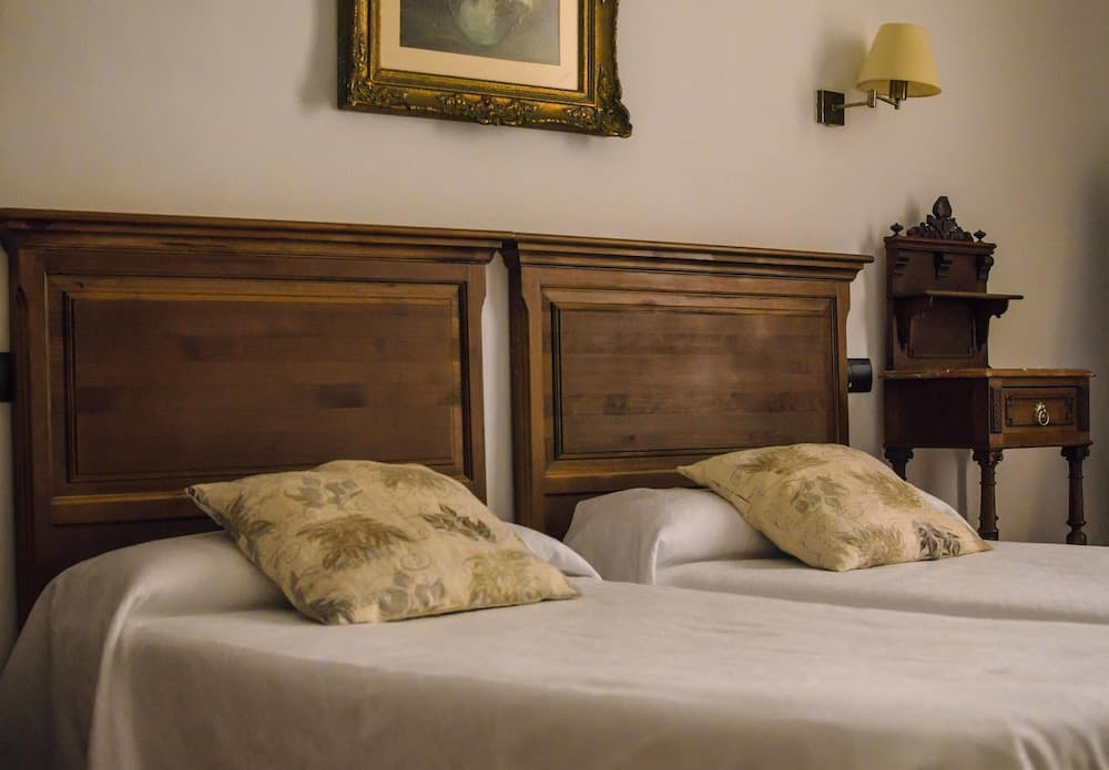 A cozy hotel room features two neatly arranged twin beds with wooden headboards, floral pillows, a vintage nightstand, and a framed artwork, ideal for travelers seeking a warm and inviting atmosphere.