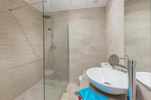 A modern bathroom with beige tiled walls, a glass-enclosed shower, and a sleek white sink creates a serene and clean atmosphere, ideal for travelers seeking comfort in accommodations.