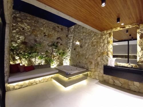 A serene indoor spa features a stone-walled room with ambient lighting, a built-in bench adorned with cushions and plants, and a modern bathtub, ideal for relaxation and wellness retreats.