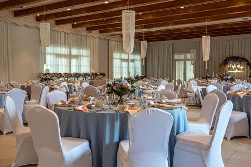 A spacious banquet hall with elegant white chairs, blue tablecloths, and large windows bathes in natural light, creating an inviting atmosphere ideal for wedding receptions or formal gatherings.