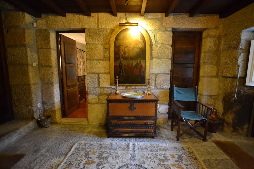 A cozy medieval-style room with stone walls, wooden beams, and a religious painting creates an intimate atmosphere, featuring a wooden chest, a folding chair, and a patterned rug, ideal for travelers seeking historical and cultural immersion.