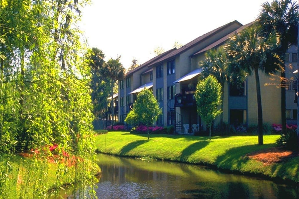 A serene waterfront apartment complex surrounded by lush greenery, palm trees, and vibrant flowers, ideal for travelers seeking a tranquil and picturesque accommodation experience.