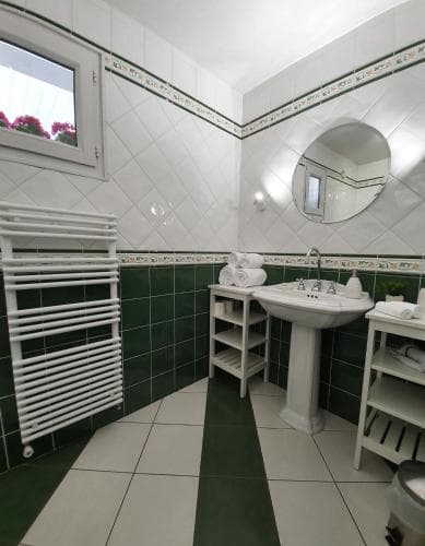 A cozy hotel bathroom features white and green tiled walls, a pedestal sink, a round mirror, neatly folded towels, and a window with a view of greenery, ideal for travelers seeking comfort and cleanliness.