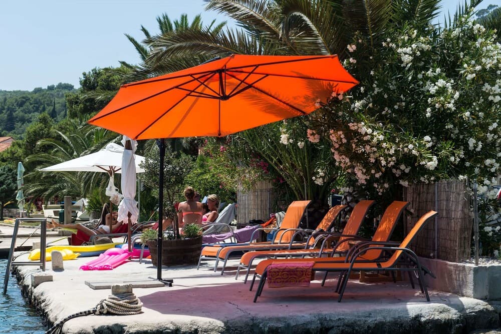 A serene waterfront scene features vibrant orange umbrellas shading lounge chairs beside lush palm trees and flowering bushes, ideal for travelers seeking relaxation in a tropical resort setting.