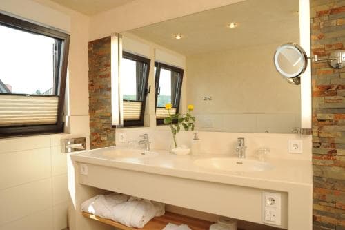 A cozy hotel bathroom features a modern double sink vanity with a large mirror, brick accent wall, fresh yellow flowers, and natural light from a window, ideal for travelers seeking comfort and style.