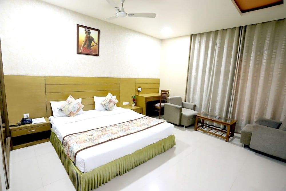 A cozy hotel room features a neatly made bed with decorative pillows, a wooden headboard, soft lighting, and a sitting area by sheer curtains, ideal for travelers seeking comfort and relaxation.
