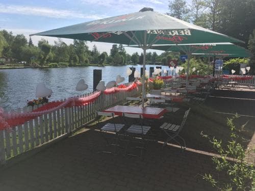 A serene lakeside café features outdoor seating under green umbrellas, surrounded by a charming white picket fence adorned with red garlands, perfect for a relaxing getaway or romantic outing.