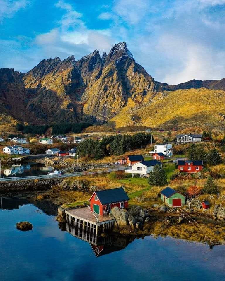 A picturesque coastal village with colorful houses nestled against dramatic mountains reflects in calm waters, creating an idyllic setting for nature lovers and photographers exploring Norway's scenic landscapes.