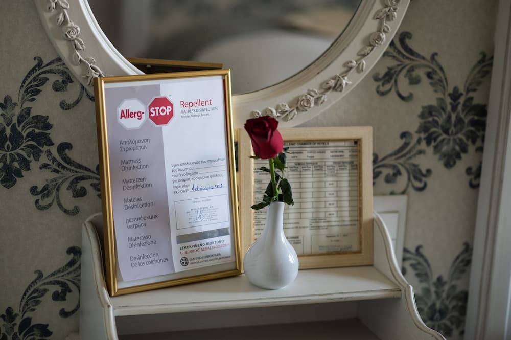 A cozy hotel room corner features an ornate mirror above a shelf holding a framed allergy stop sign, a vase with a single red rose, and a certificate, evoking a welcoming and hygienic atmosphere ideal for travelers seeking comfort and cleanliness.