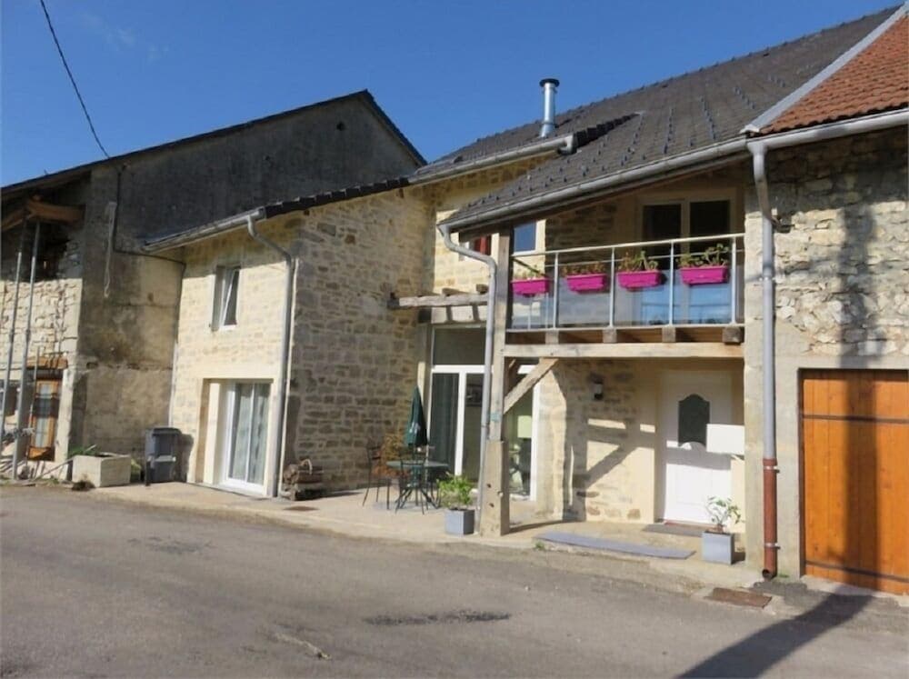 A charming stone cottage with a balcony adorned with pink flower pots stands in a quaint village setting, perfect for a serene countryside getaway.