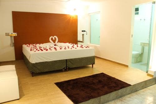 A cozy hotel room features a neatly arranged bed adorned with rose petals and a heart-shaped pillow, set against a warm wooden backdrop, complemented by a modern bathroom and inviting ambiance ideal for romantic getaways.