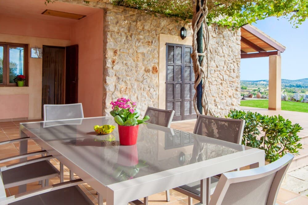 A serene outdoor dining area features a modern glass table set against a rustic stone wall, surrounded by lush greenery and overlooking a tranquil landscape, ideal for a relaxing vacation retreat.
