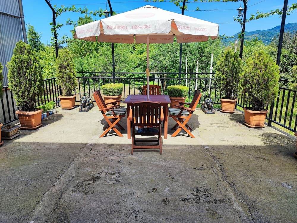 A serene outdoor patio features wooden furniture under a large umbrella, surrounded by lush greenery and potted plants, ideal for travelers seeking a peaceful retreat in a scenic mountainous area.