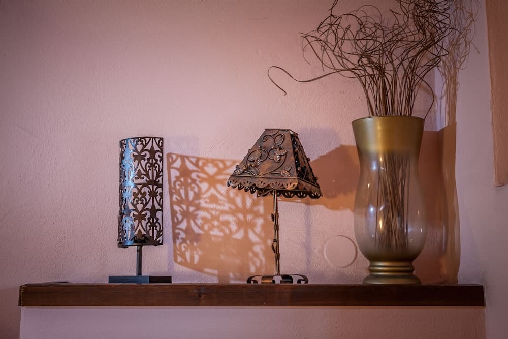 A cozy indoor setting features a wooden shelf adorned with an ornate lamp casting intricate shadows, a decorative vase with dried branches, and a metallic lantern, ideal for travelers seeking warm and artistic accommodations.