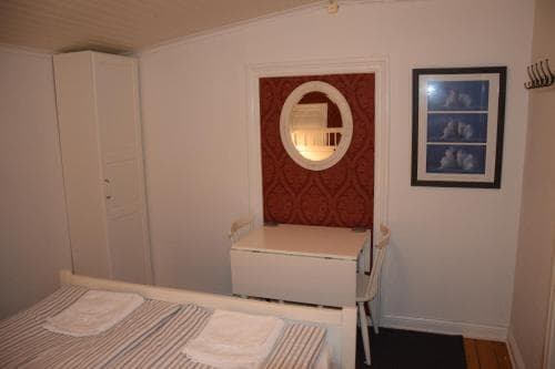 A cozy bedroom with a neatly made bed, white furniture, a red patterned wall panel with an oval mirror, and framed artwork creates a serene atmosphere ideal for travelers seeking a comfortable and intimate accommodation experience.