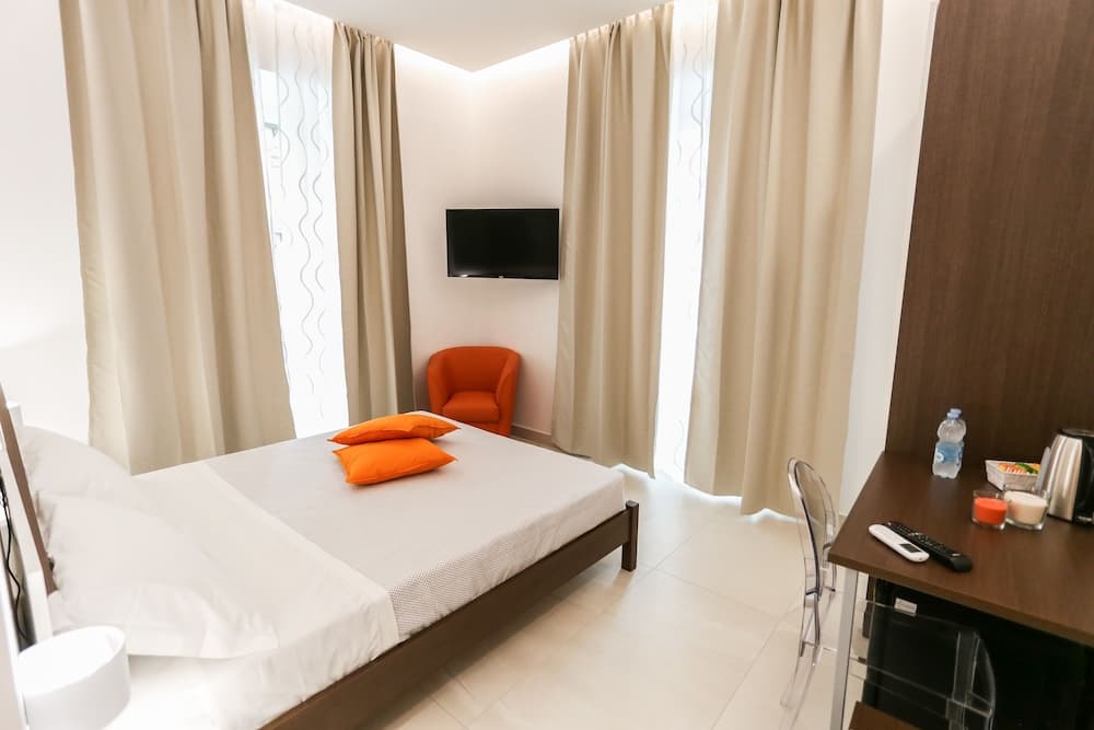 A cozy hotel room features a neatly made bed with orange pillows, a bright orange armchair, a mounted TV, and a desk with amenities, ideal for travelers seeking comfort and convenience.