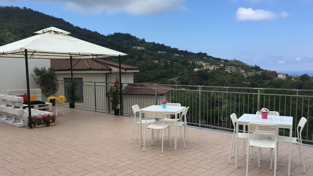 A serene rooftop terrace with white tables, chairs, and an umbrella overlooks lush green hills dotted with houses under a clear blue sky, ideal for a relaxing vacation getaway.