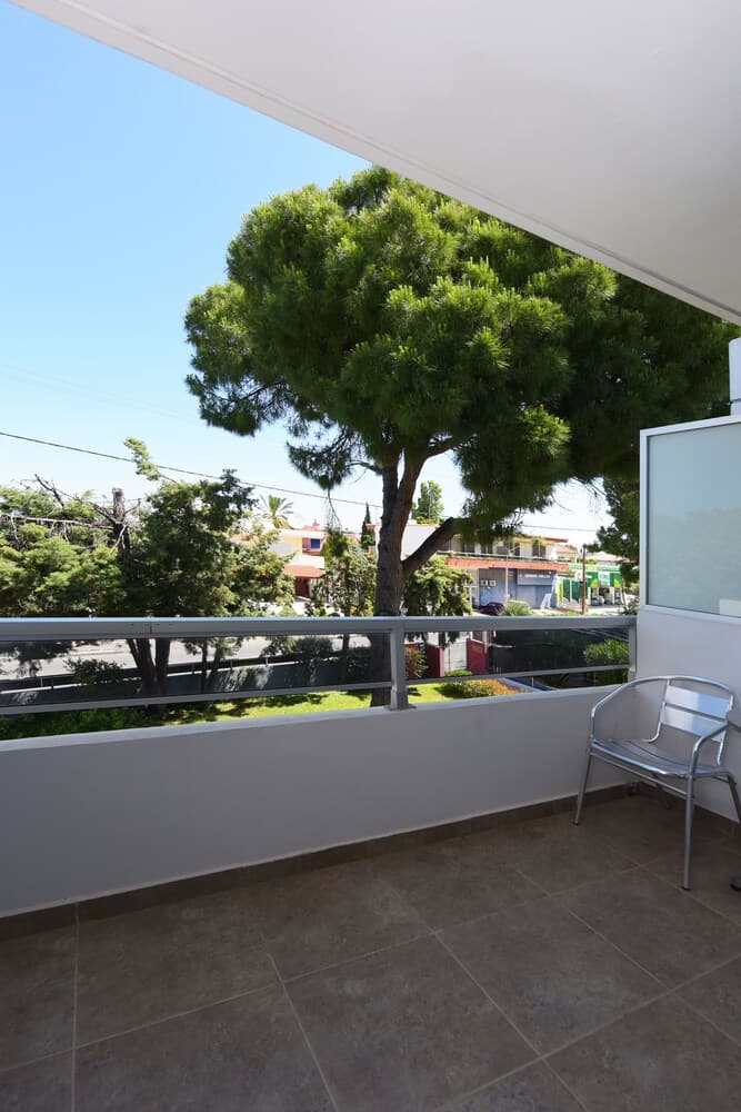 A serene balcony with a metal chair overlooks lush greenery and distant buildings under a clear blue sky, ideal for travelers seeking a peaceful retreat in an urban setting.
