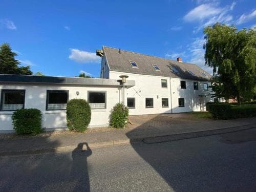 A serene suburban street features a white building with a sloped roof under a clear blue sky, surrounded by lush greenery, ideal for travelers seeking a peaceful accommodation experience.