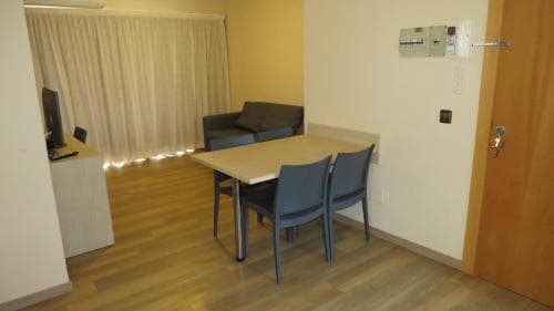 A cozy hotel room features a wooden dining table with chairs, a sofa, and a desk by the window, creating a warm and functional space ideal for travelers seeking comfort and convenience.