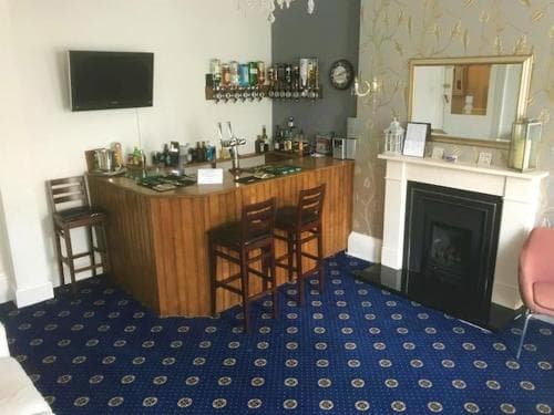 A cozy hotel lounge features a small bar with stools, a fireplace adorned with a mirror, and a blue patterned carpet, creating an inviting atmosphere for travelers seeking relaxation.