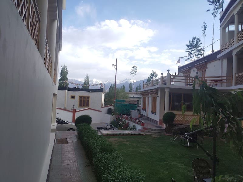 A serene courtyard at Hotel Ladakh Inn in Leh showcases lush greenery, traditional architecture, and distant snow-capped mountains under a bright sky, ideal for travelers seeking a peaceful Himalayan retreat.