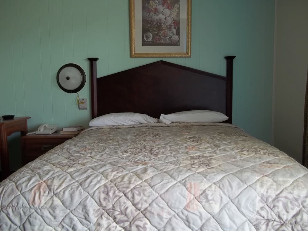 A cozy bedroom with teal walls, a dark wooden bed adorned with a quilted floral comforter, a framed floral artwork above the headboard, and a bedside table featuring a phone and books, ideal for travelers seeking a serene and comfortable stay.