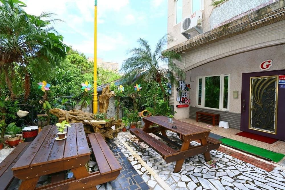 A cozy tropical courtyard features wooden picnic tables surrounded by lush greenery, colorful pinwheels, and mosaic-tiled pathways, ideal for a relaxing getaway or family vacation spot.