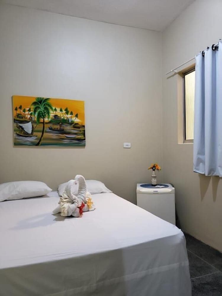 A cozy hotel room features a neatly made bed adorned with a towel swan, a tropical painting on the wall, and a small table with flowers by the window, ideal for travelers seeking a tranquil and inviting accommodation.