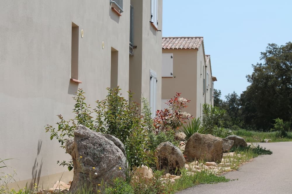 A serene residential street lined with beige houses featuring terracotta roofs, lush greenery, and large decorative rocks creates an inviting atmosphere ideal for a peaceful vacation getaway.