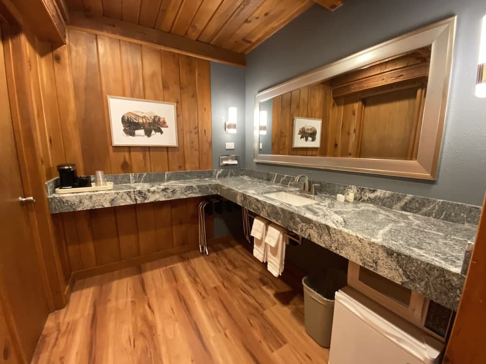 A cozy cabin-style bathroom features wood-paneled walls, a granite countertop, a large mirror, and a bear-themed artwork, ideal for travelers seeking rustic charm in their accommodations.