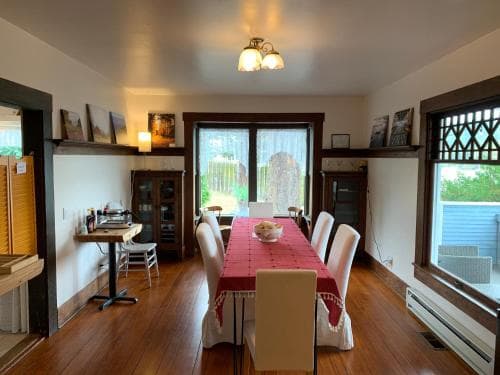 A cozy dining room with hardwood floors, a red tablecloth-covered table, white chairs, and large windows offering a serene view, ideal for travelers seeking a warm and inviting accommodation experience.
