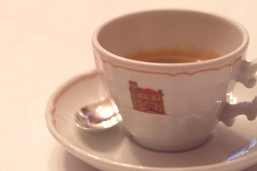 A cozy café ambiance is captured with a close-up of an espresso cup adorned with a building illustration, resting on a saucer with a spoon, ideal for travelers seeking authentic coffee experiences.