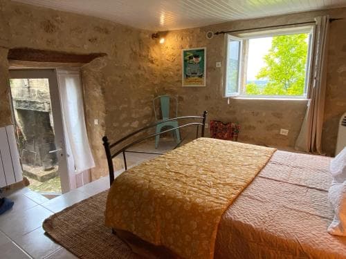 A cozy stone-walled bedroom with a rustic charm, featuring a neatly made bed, a window with lush greenery outside, and a quaint chair, ideal for travelers seeking a serene countryside retreat.