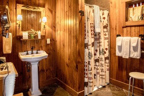 A cozy cabin bathroom with warm wooden walls, a rustic shower curtain featuring forest motifs, a vintage pedestal sink, and soft lighting creates an inviting retreat perfect for travelers seeking a serene getaway experience.