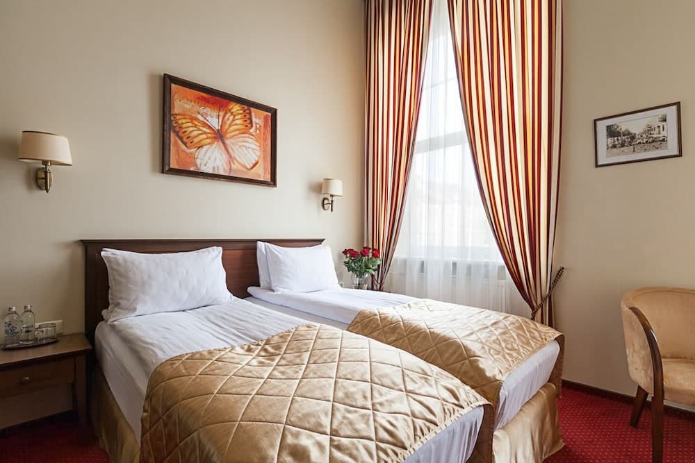 A cozy hotel room features twin beds with golden quilts, striped curtains, wall-mounted lamps, and a butterfly painting, creating a warm and inviting atmosphere ideal for travelers seeking comfort.