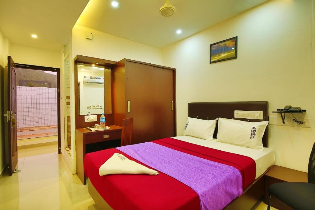 A cozy hotel room features a neatly made bed with vibrant red and purple bedding, a small desk with amenities, and an open door leading to a balcony, ideal for travelers seeking comfort and convenience.