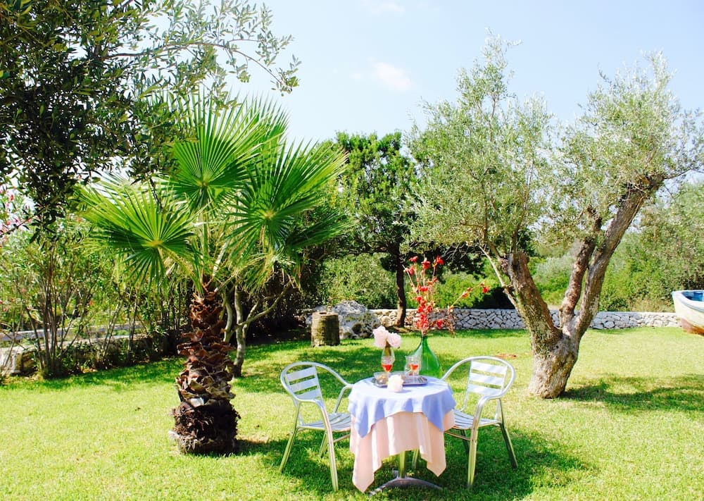 A serene garden setting features a cozy table draped in pastel linens surrounded by lush olive trees and a vibrant palm, ideal for a tranquil outdoor dining experience.