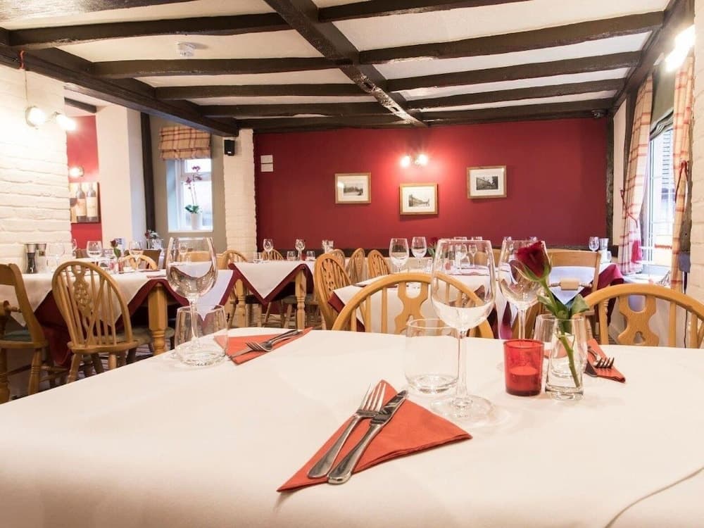 A cozy restaurant with rustic wooden beams, red walls adorned with framed pictures, and tables set with white linens, orange napkins, and elegant glassware, perfect for a romantic dinner or intimate gathering.