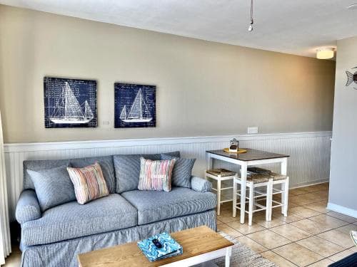 A cozy living room with nautical-themed decor, featuring sailboat artwork, a comfortable sofa, and a small dining area, creates an inviting atmosphere ideal for a relaxing beachside vacation rental.