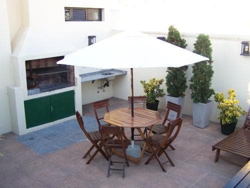 A cozy outdoor patio features a wooden table and chairs under a white umbrella, surrounded by potted plants and a built-in barbecue, ideal for relaxing or hosting small gatherings in a serene urban setting.