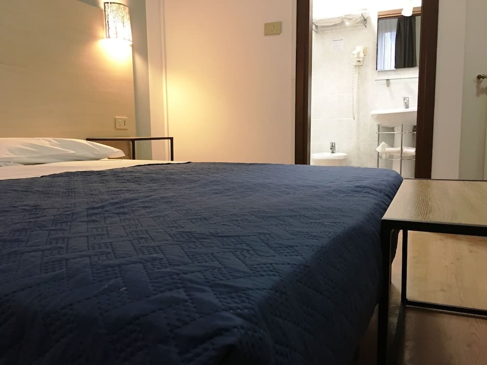 A cozy hotel room features a neatly made bed with a blue quilt, a warm wall lamp, and an open door leading to a clean bathroom, ideal for travelers seeking comfort and convenience.