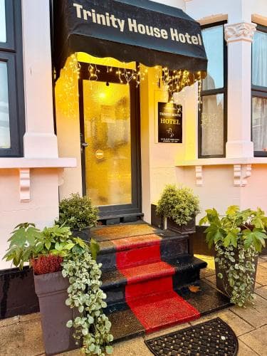 The warmly lit entrance of Trinity House Hotel, adorned with festive lights and lush potted plants, invites travelers to a cozy stay in an urban setting.