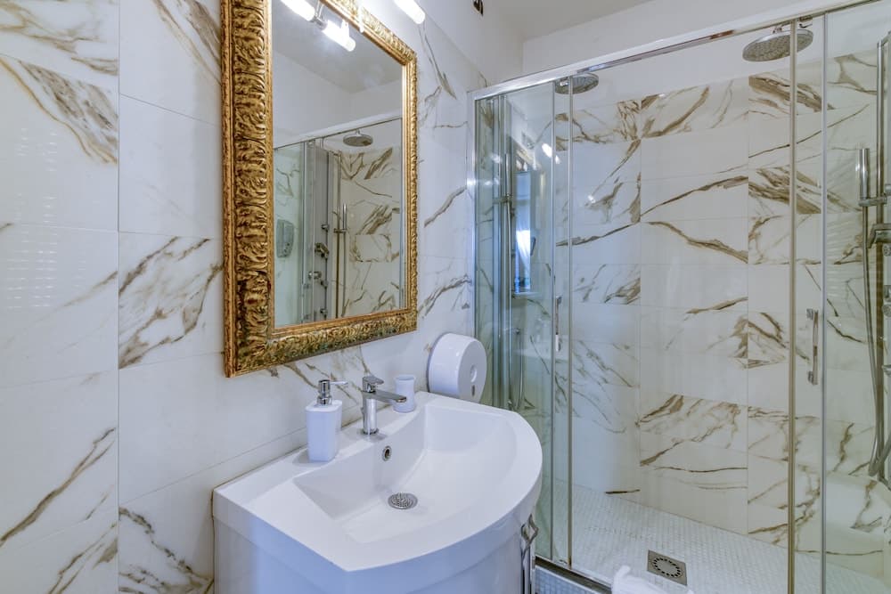 A luxurious bathroom with marble tiles, a sleek glass shower enclosure, and an ornate gold-framed mirror exudes elegance, ideal for travelers seeking premium accommodations.