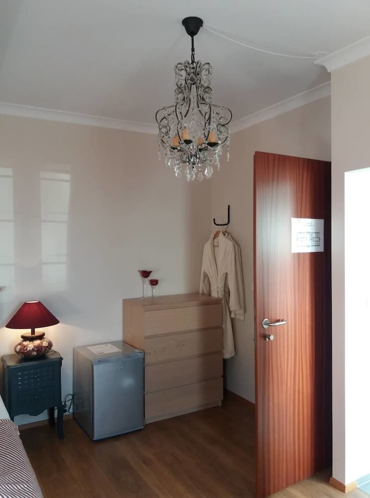 A cozy hotel room features a chandelier, wooden dresser, mini fridge, and robe hanging by the door, creating an inviting atmosphere ideal for travelers seeking comfort and convenience.