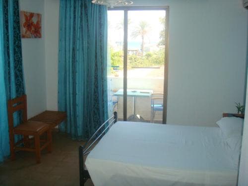 A cozy hotel room with turquoise curtains frames a serene balcony view of palm trees and the sea, ideal for travelers seeking relaxation near the beach.