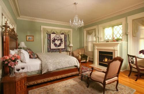A cozy, elegantly furnished bedroom with soft green walls, a fireplace, chandelier, and floral accents creates an inviting atmosphere perfect for a romantic getaway or luxurious staycation.